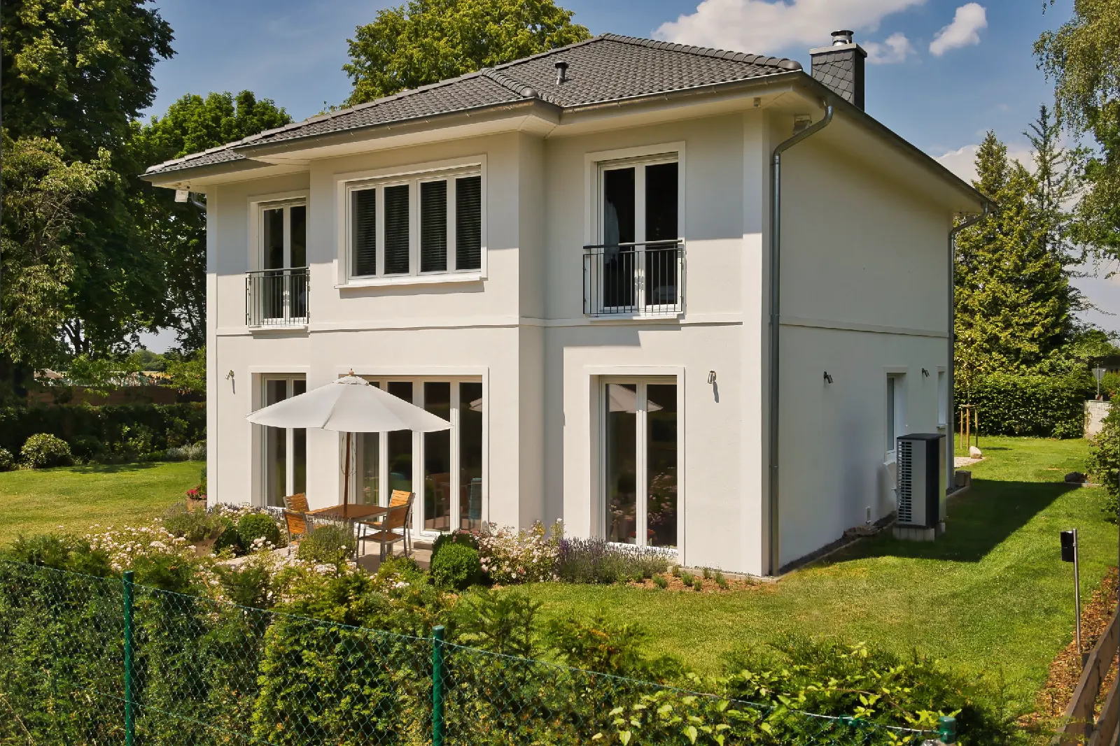 Ein zweigeschossiges weißes Einfamilienhaus steht in einem begrünten Garten.