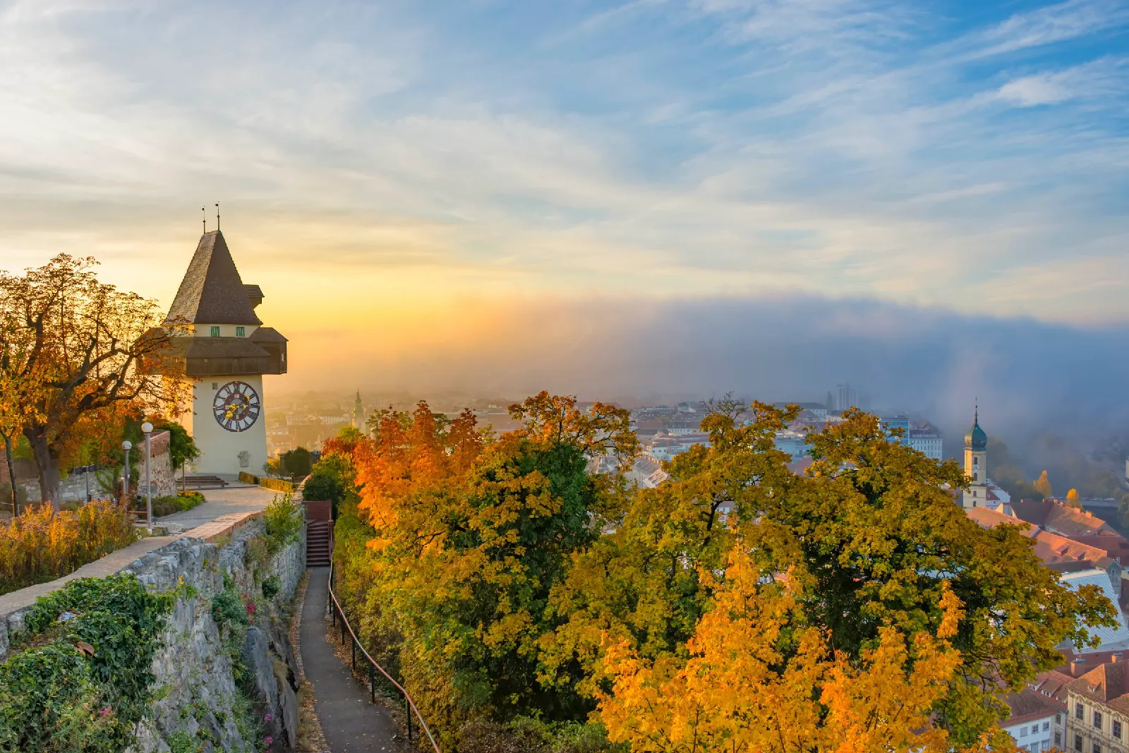 Grazer Uhrturm und Dächer der Stadt zwischen herbstlichen Bäumen und aufziehendem Nebel